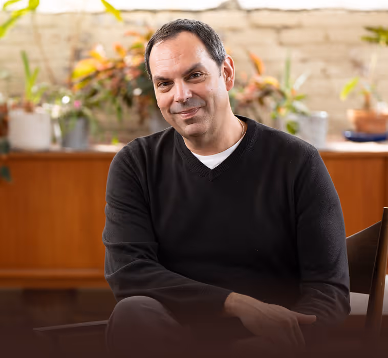 Man with short dark hair wearing a black sweater and white shirt, sitting indoors with various plants and a wooden cabinet in the background.