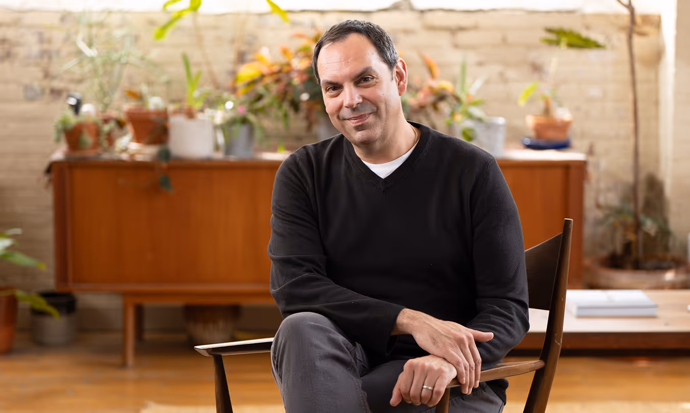 Smiling man with short dark hair wearing a black sweater and gray pants, sitting casually on a wooden chair in a bright room with a wooden sideboard and several potted plants in the background.