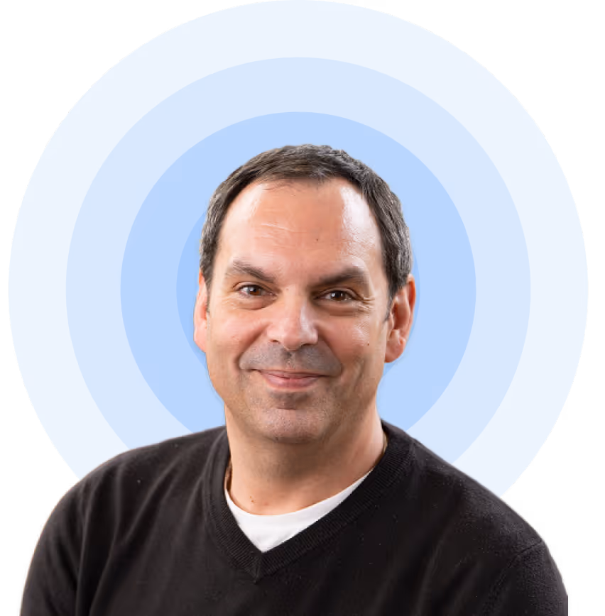 Smiling middle-aged man with short dark hair wearing a black sweater over a white shirt, against a light blue circular background.