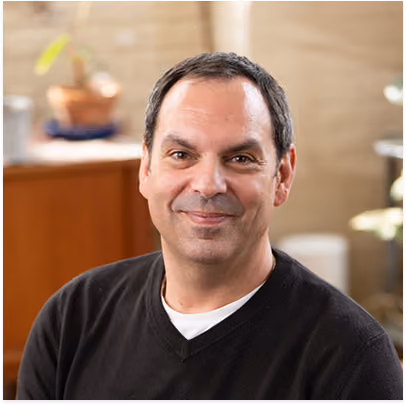 Middle-aged man with short dark hair wearing a black V-neck sweater over a white shirt, smiling in a warmly lit indoor setting.