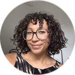 Woman with curly dark hair wearing large glasses and a sleeveless patterned top, smiling indoors against a light gray wall.