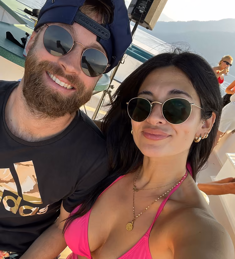 Smiling couple wearing sunglasses taking a selfie outdoors near a body of water with mountains in the background; the man has a beard and wears a backwards cap and black Adidas shirt while the woman wears a pink bikini top and gold jewelry.