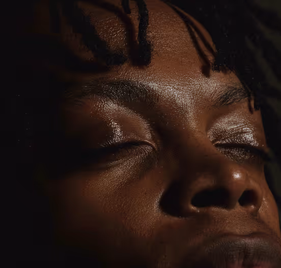 Close-up of a person with dark skin and closed eyes, with textured skin and a few strands of hair falling gently over the forehead.