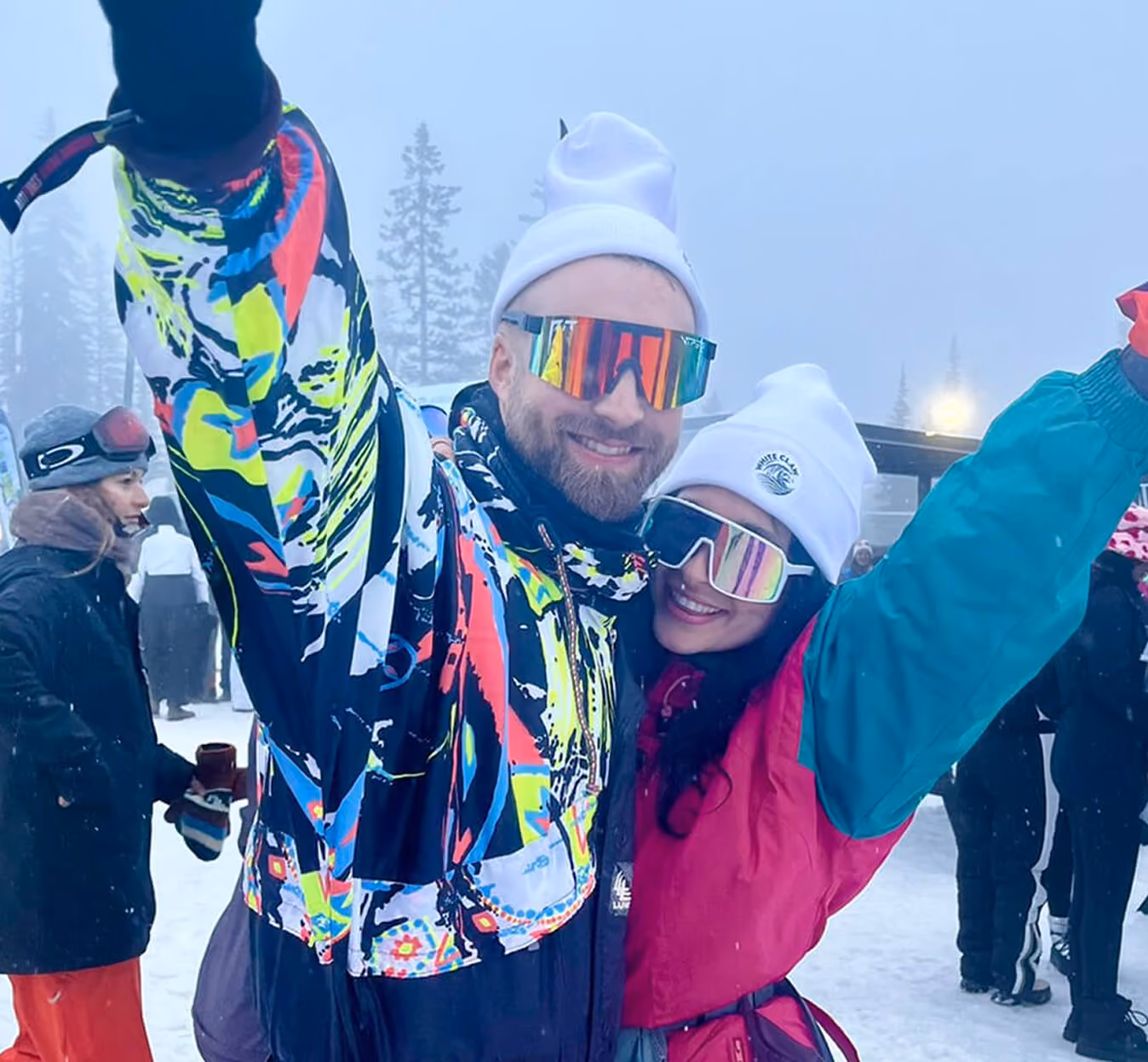 A smiling couple wearing colorful ski jackets, white winter hats, and reflective ski goggles posing with raised arms in a snowy outdoor setting with other people and trees in the background.