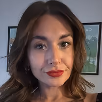 Close-up portrait of a woman with shoulder-length brown hair, wearing red lipstick and earrings, with framed art on the wall in the background.